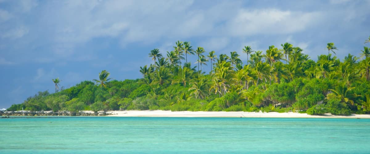 Une plage tropicale avec une eau turquoise et limpide, du sable blanc et de luxuriants palmiers verdoyants, le tout sous un ciel partiellement nuageux.