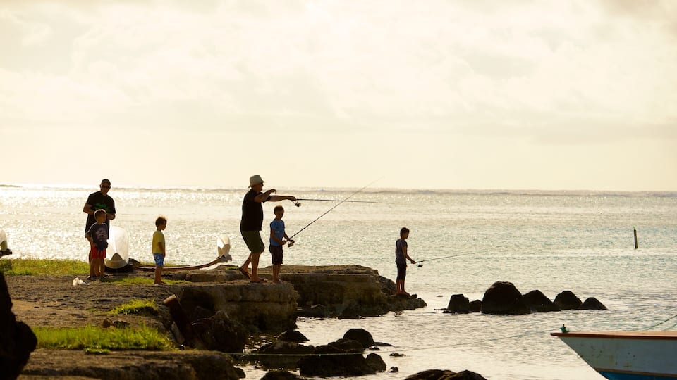 A group of people fishing by a rocky shoreline with calm water and a boat in the background.