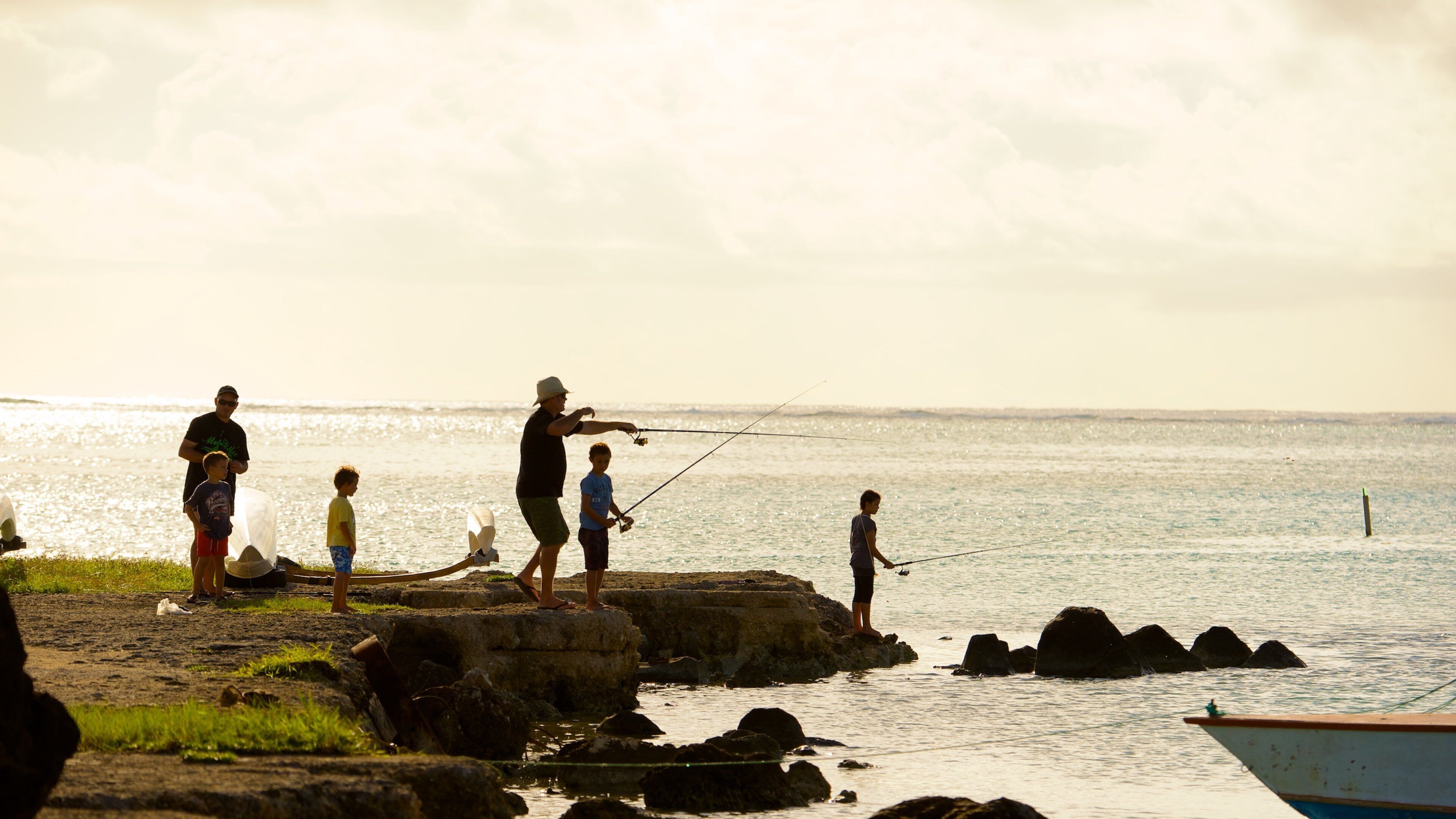 A group of people fishing by a rocky shoreline with calm water and a boat in the background.