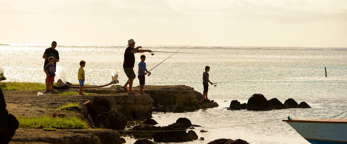 A group of people fishing by a rocky shoreline with calm water and a boat in the background.