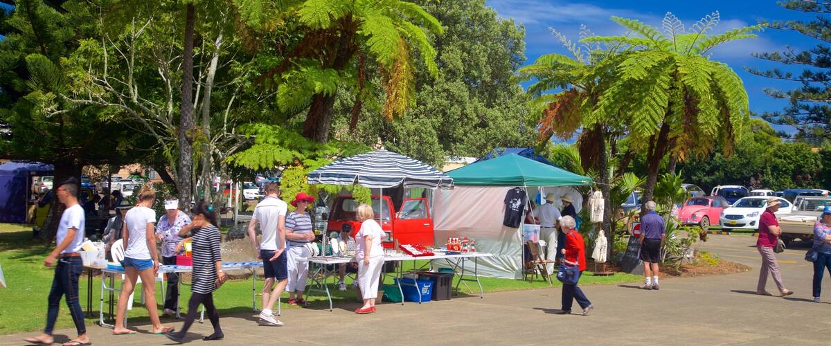A sunny outdoor market with people walking around, tents, and parked cars.