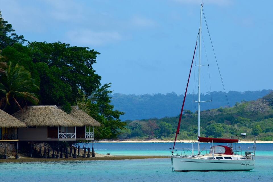 A sailboat anchored near a thatched-roof hut on a clear turquoise beach, surrounded by lush greenery and a distant mountain range.