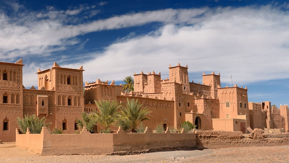 Un grand bâtiment en briques de terre, aux motifs complexes, doté de nombreuses tours et d’arcades, entouré de palmiers et surplombé d’un ciel bleu azur.