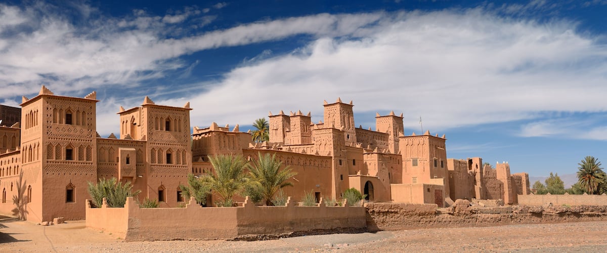 Un edificio grande de adobe, ricamente decorado, con múltiples torres y arcos, rodeado de palmeras y un cielo azul despejado.