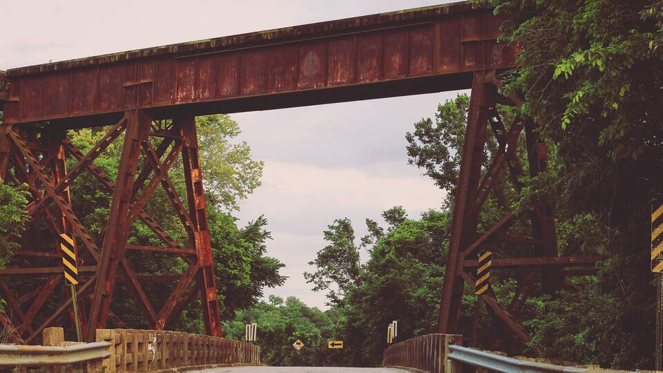 A rusted railway bridge with a road underneath, surrounded by dense trees.