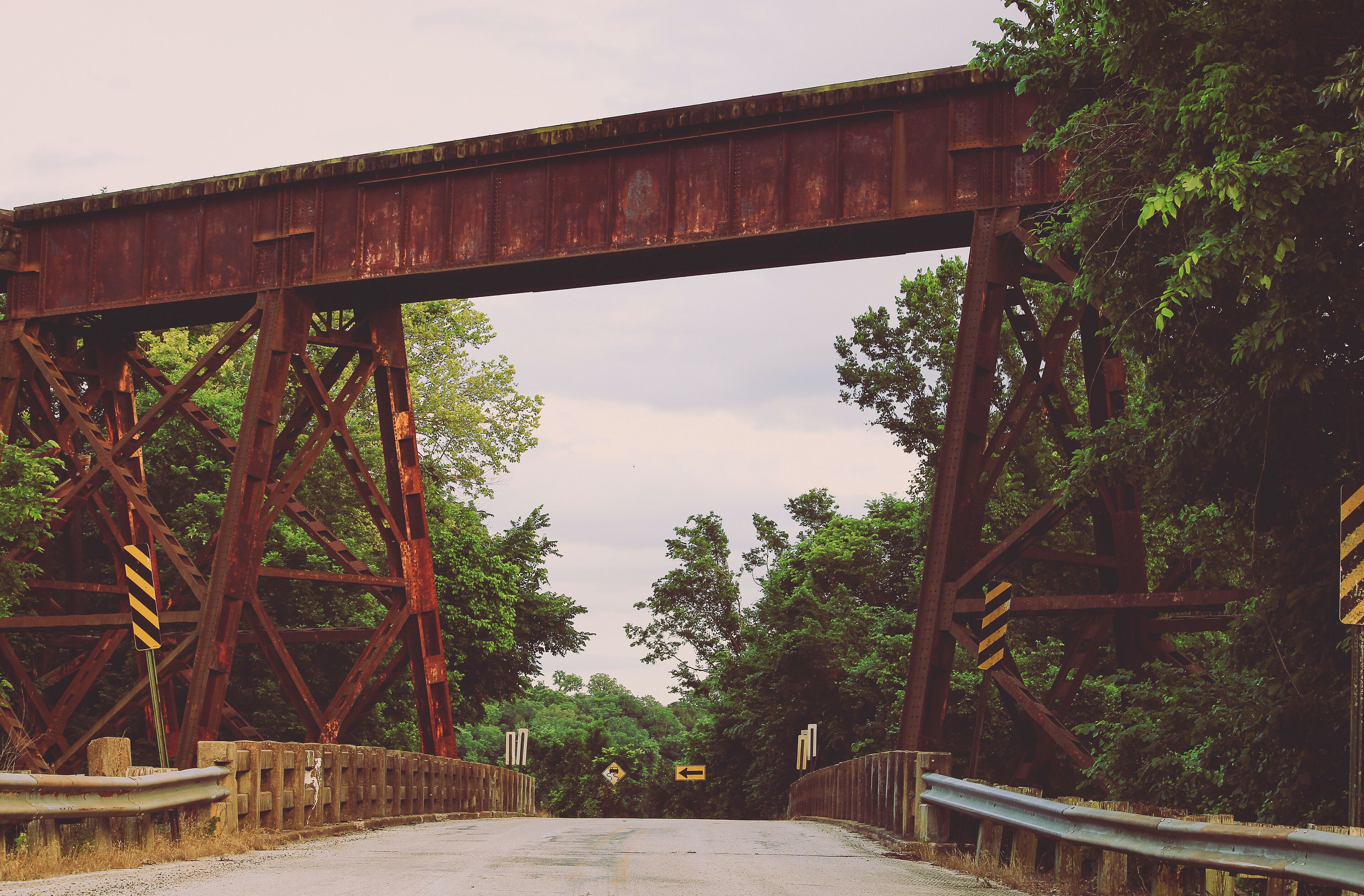 A rusted railway bridge with a road underneath, surrounded by dense trees.