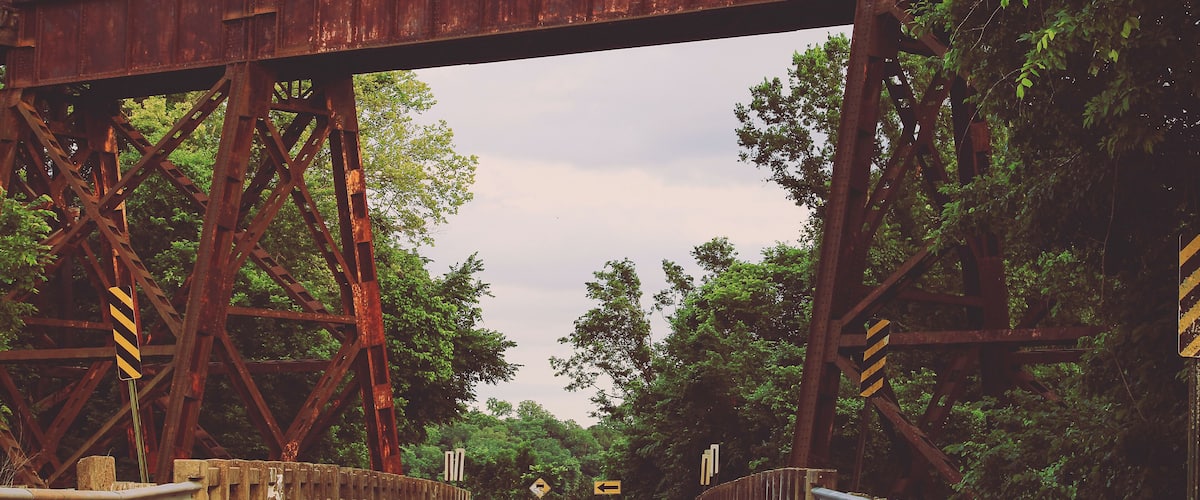 A rusted railway bridge with a road underneath, surrounded by dense trees.