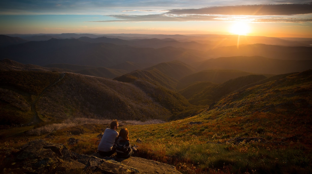 Una pareja sentada sobre una roca, con vista a un valle y el atardecer de fondo.