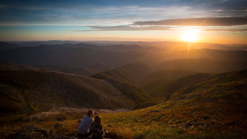 A couple sitting on a rock overlooking a valley with a sunset in the background.