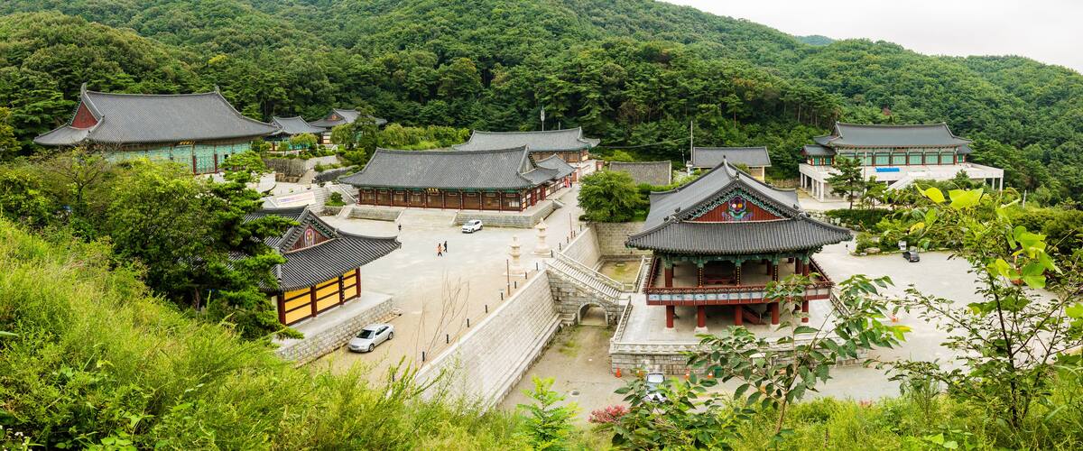 A traditional Korean temple complex with multiple buildings, surrounded by lush greenery and hills.