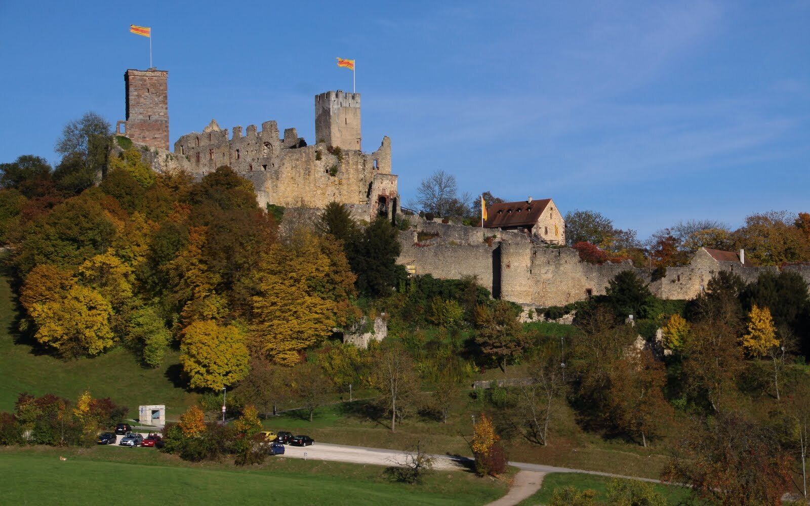 A historic castle with two towers, surrounded by autumn-colored trees and a small building.