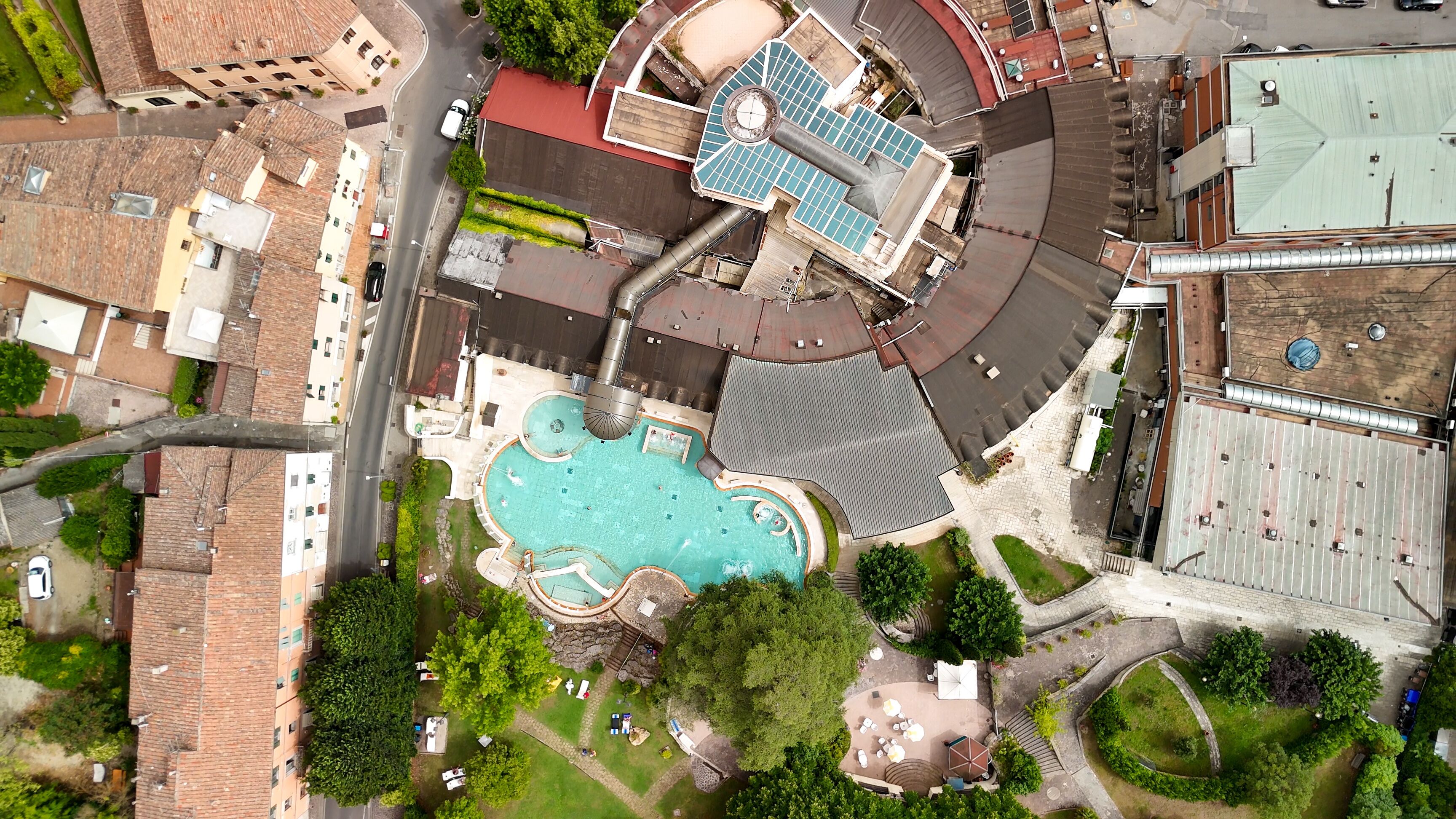 An aerial view of a residential area with a swimming pool, a large building with a red roof, and surrounding greenery.