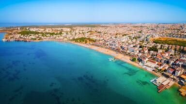 An aerial view of a coastal city with a beach, dense urban area, and clear blue waters.