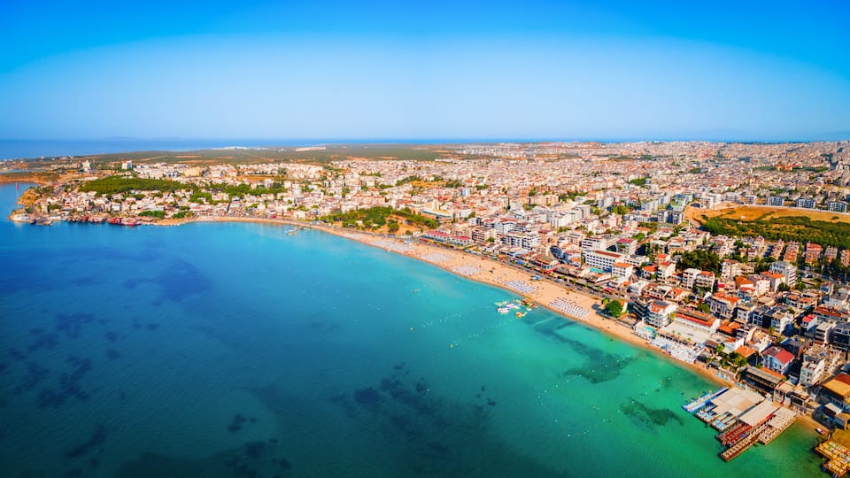 An aerial view of a coastal city with a beach, dense urban area, and clear blue waters.