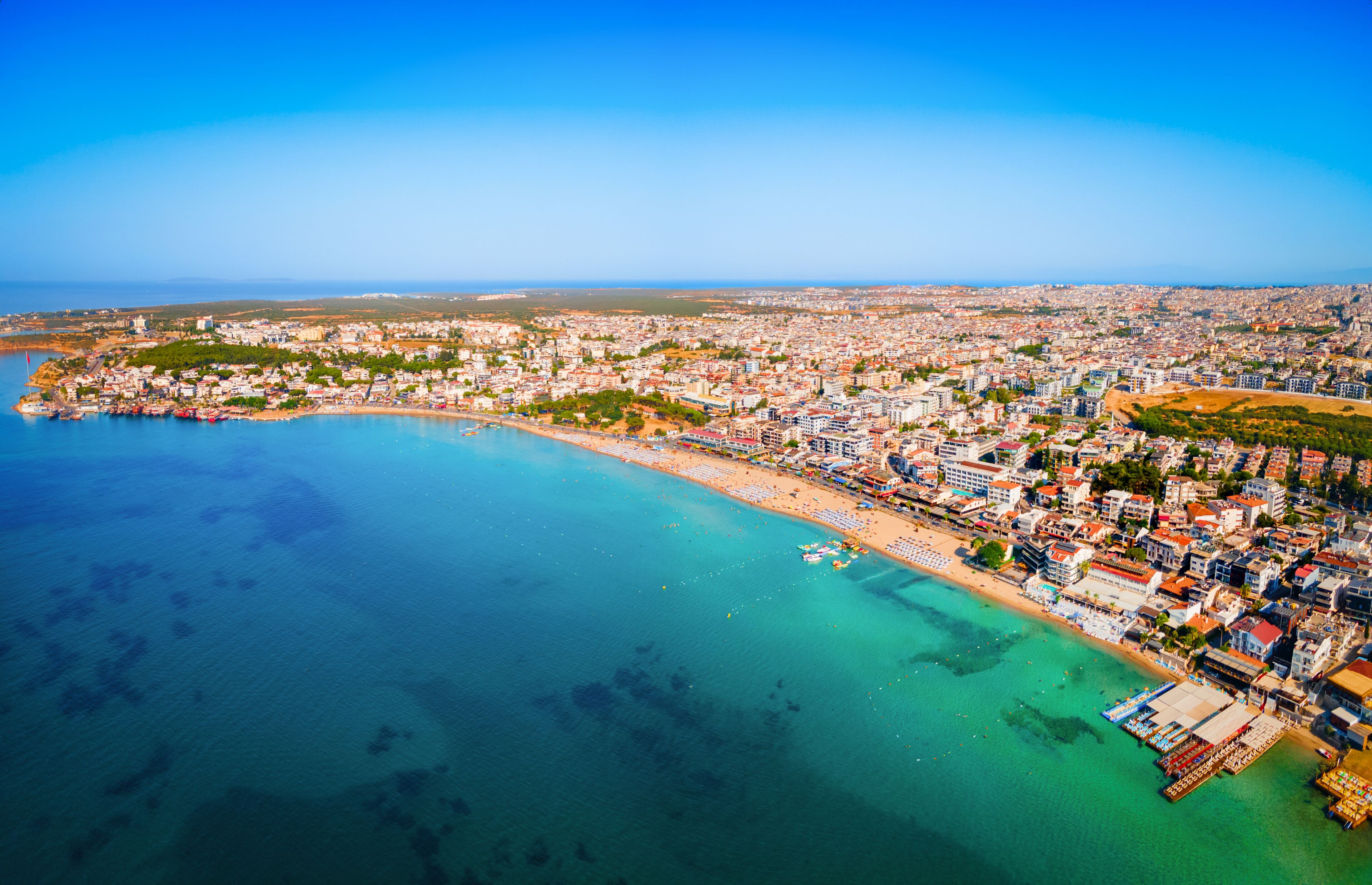 An aerial view of a coastal city with a beach, dense urban area, and clear blue waters.