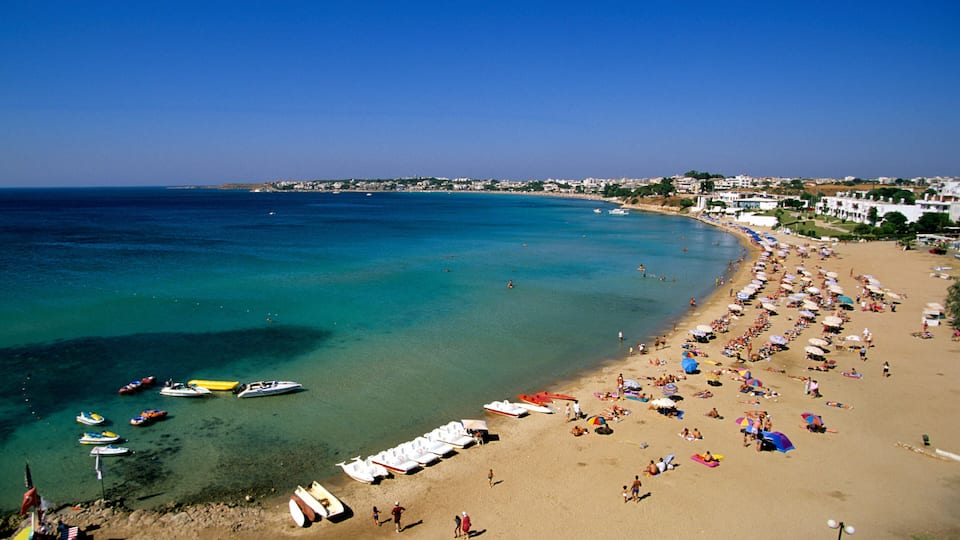 A beach with clear turquoise water, numerous boats, and a crowded sandy shore with umbrellas.