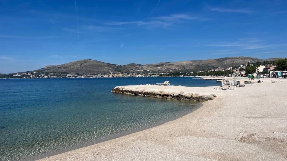 A clear coastal beach with a rocky breakwater, calm sea, and a backdrop of hills and buildings.