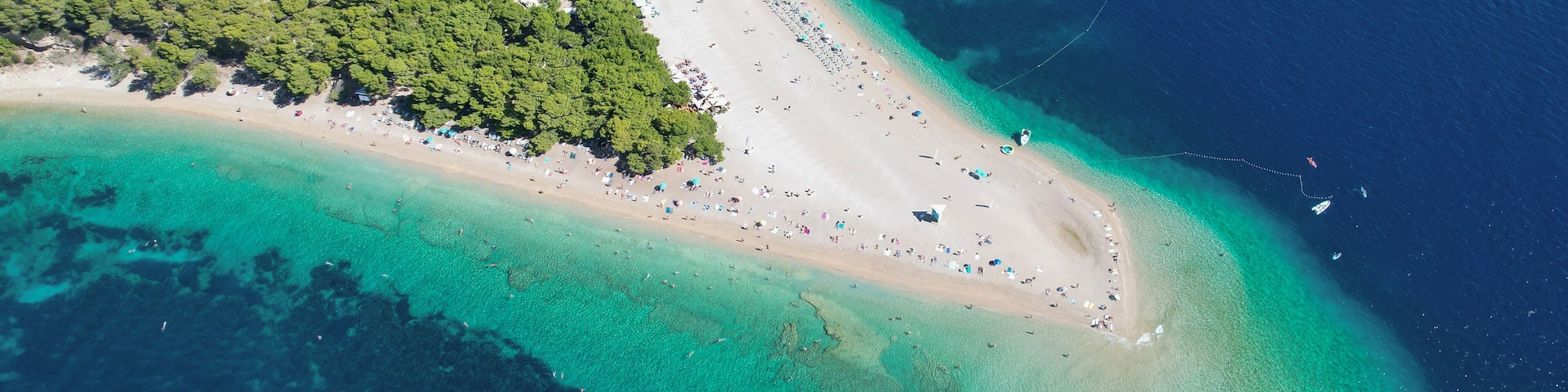 An aerial view of a coastal area with a sandy beach, clear turquoise waters, and dense green forest.