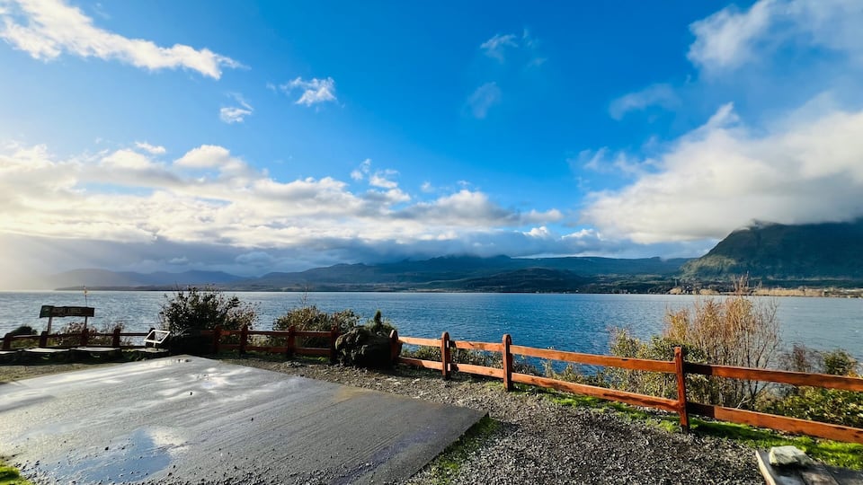 A lakeside view with a wooden fence, a paved area, and mountains in the background.