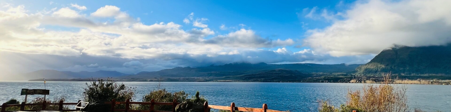 Vista de un lago con una valla de madera, un área pavimentada y montañas al fondo.