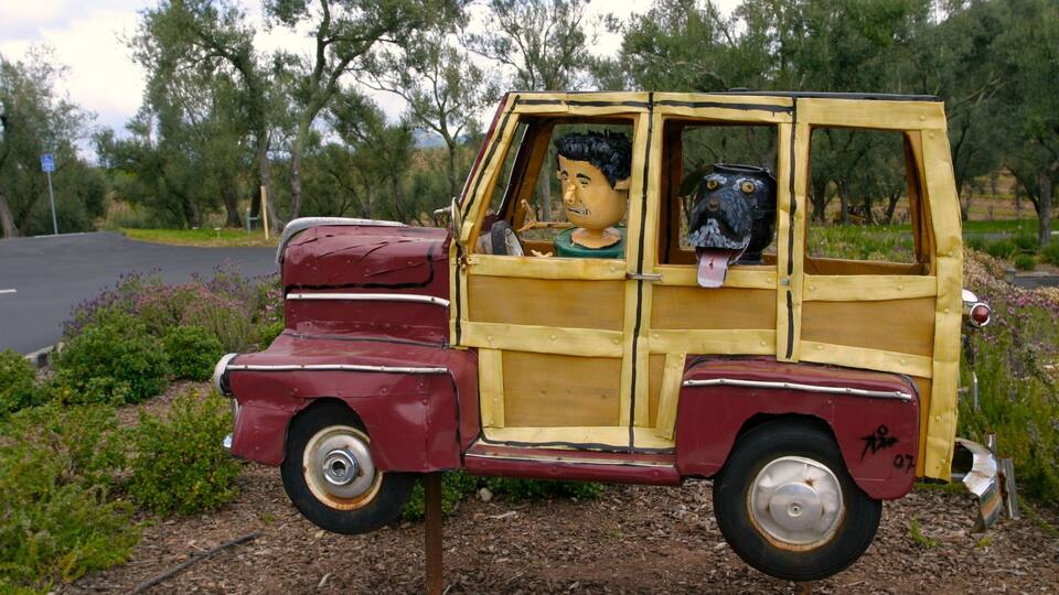 A rustic wooden car with a large face on the front and another on the back, parked on a dirt path with trees in the background.