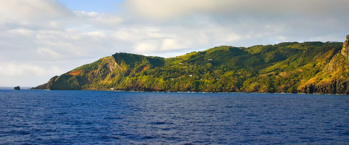 A coastal landscape with a clear sky, a calm sea, and green hills with scattered buildings.