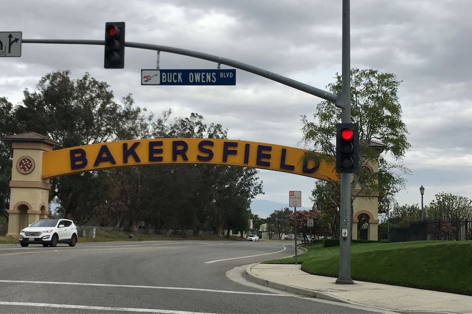 A street sign for Buck Owens Blvd and Bakersfield, with a white SUV on the road.