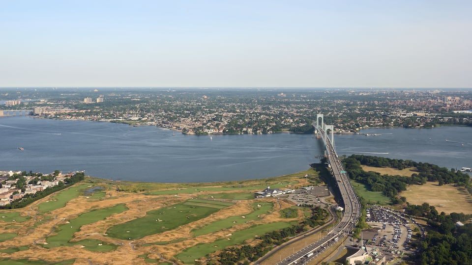 A coastal cityscape with a prominent bridge spanning a wide river, surrounded by urban areas and green spaces.