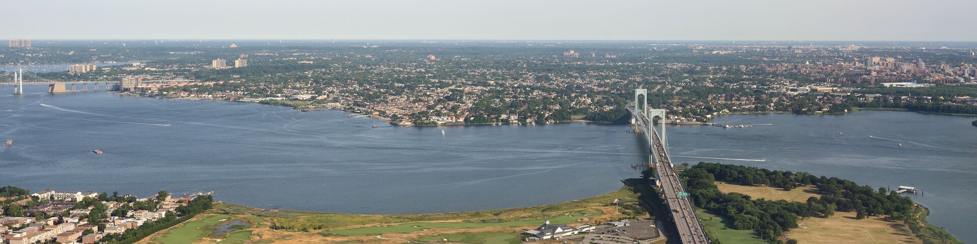 Paisagem costeira urbana com uma ponte imponente sobre um rio largo, rodeada por áreas urbanas e espaços verdes.