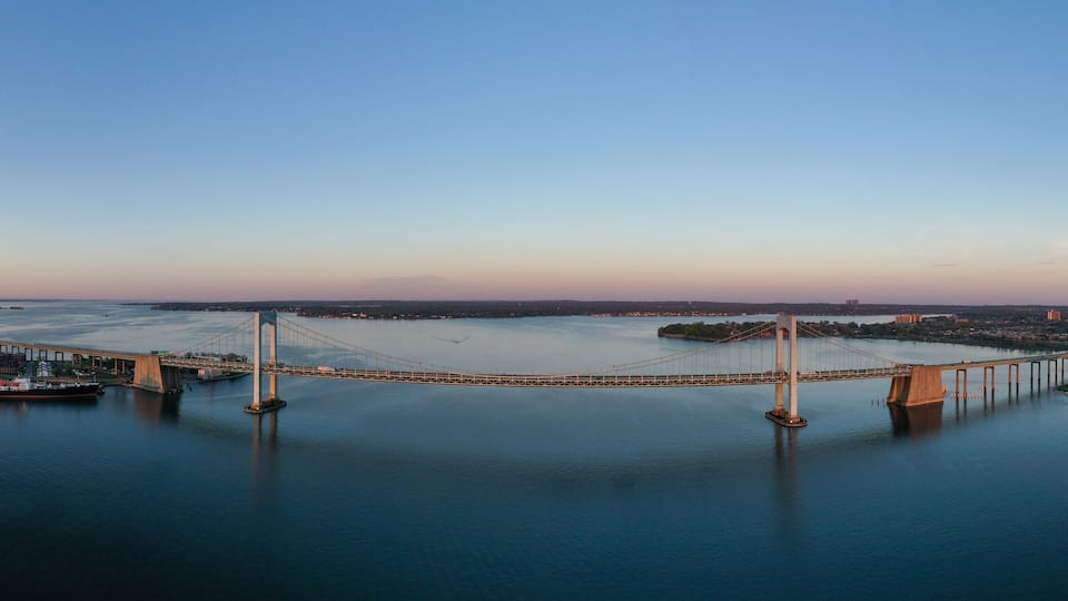 A large suspension bridge spanning a wide river, surrounded by urban development and a distant shoreline.