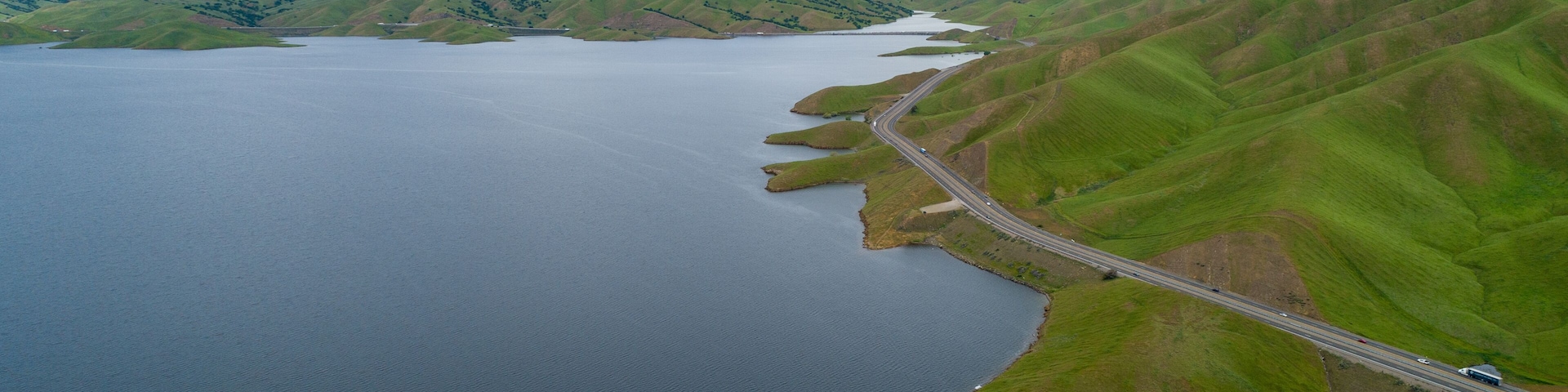 Um lago rodeado por colinas verdes, uma estrada sinuosa e um céu nublado.