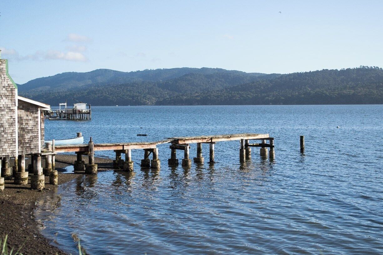 A wooden pier extending into a calm lake, with a small building on the left and mountains in the background.