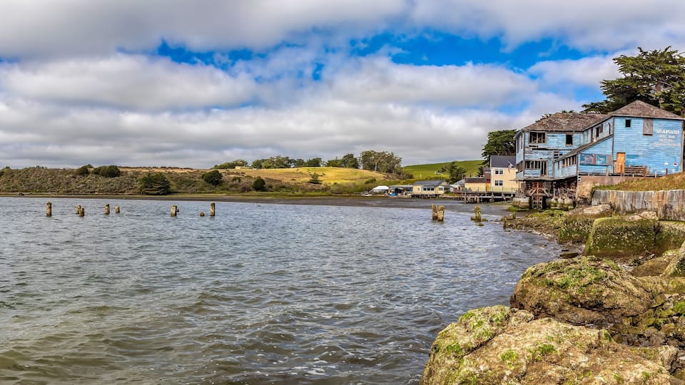 A coastal scene with a rocky shoreline, a blue building with a sign reading "SEAFOOD," and a distant view of a hill with sparse vegetation.