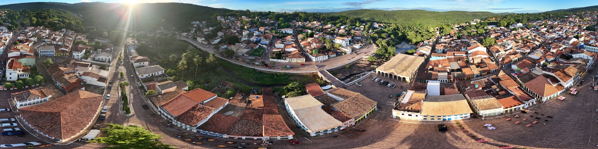 A panoramic view of a historic town with terracotta-roofed buildings, a central square, and surrounding hills.