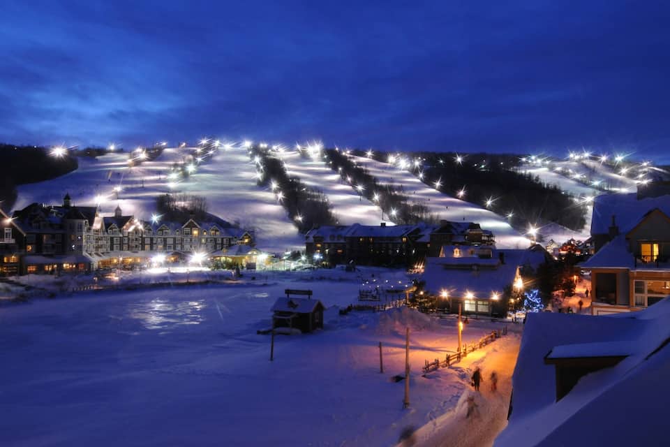 Estación de esquí nevada de noche con edificios iluminados y pistas de esquí.