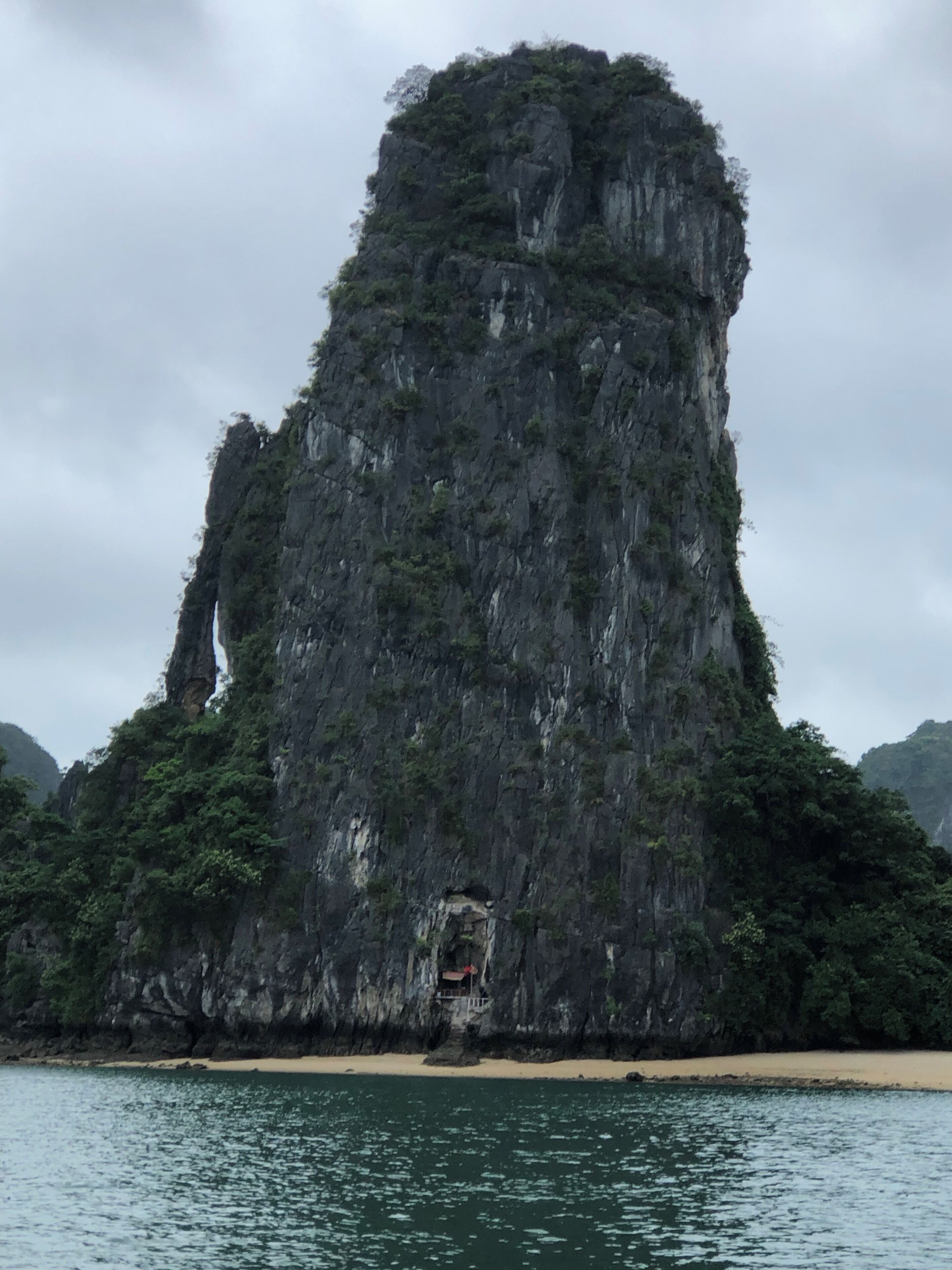 A large limestone karst formation with a cave entrance at its base, surrounded by lush greenery and a sandy beach.