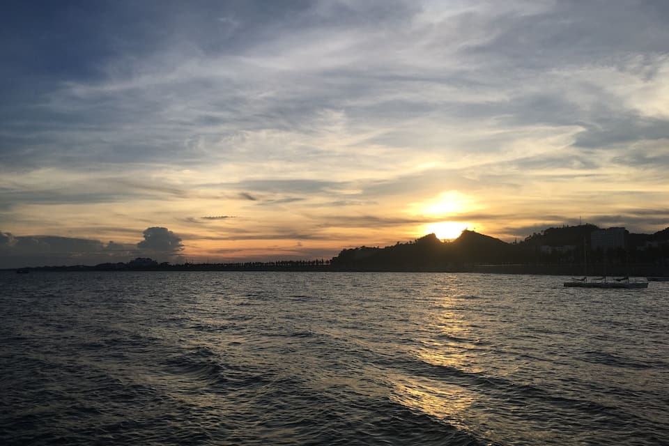 A sunset over a body of water with a silhouette of a hill and buildings in the background.