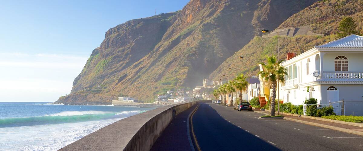 A coastal road with a cliff, ocean waves, and white buildings with palm trees.