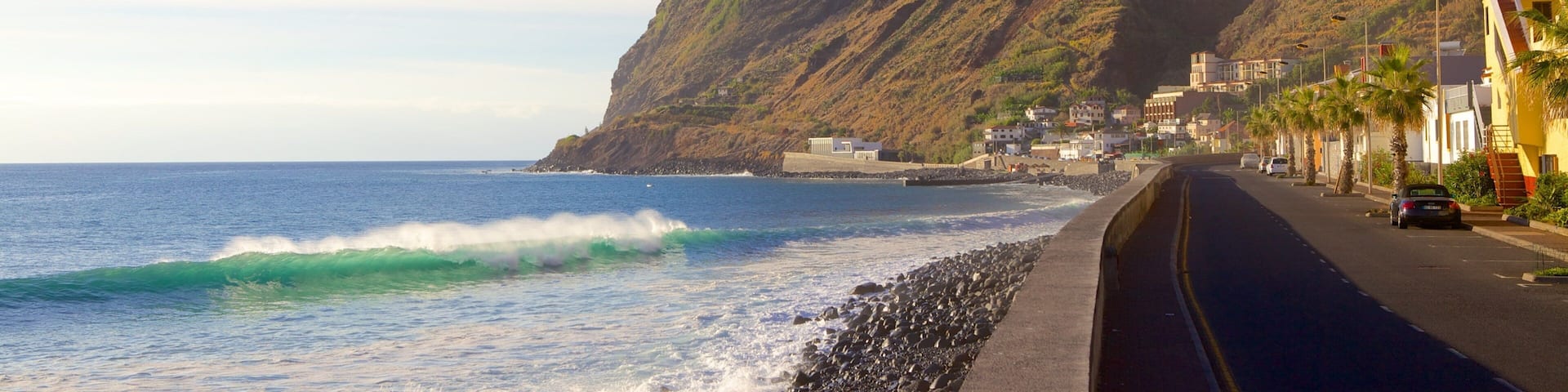 A coastal road with a steep cliff, ocean waves, and a row of buildings along the seaside.