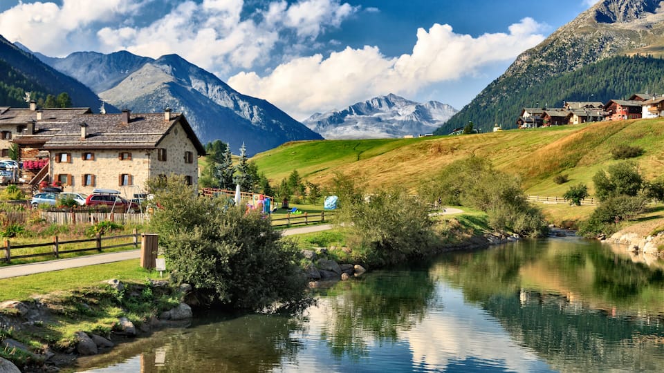 Calm River Flowing Past Quaint Buildings Village of Livigno in Foothills of Italian Alps on Sunny Day Under Blue Sky