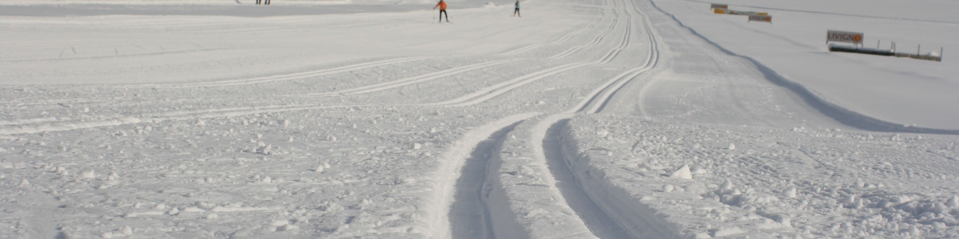 Una pista de esquí nevada con huellas, algunos esquiadores y edificios al fondo.