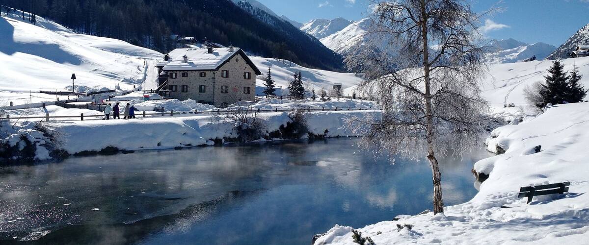 A snow-covered landscape with a river, a wooden house, and mountains in the background.