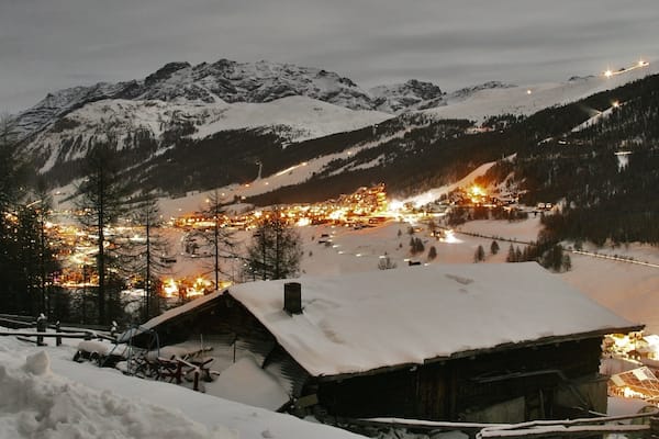 A snow-covered mountain village at night with illuminated buildings and a ski slope.