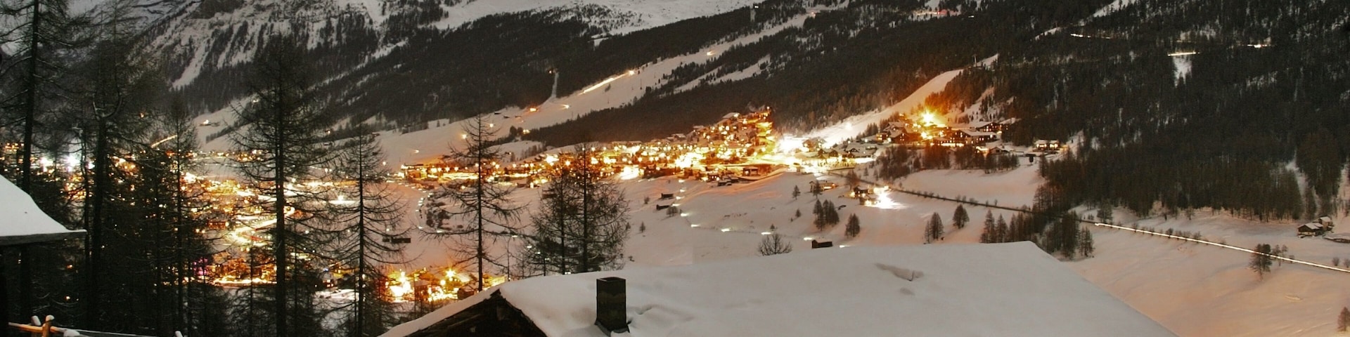 A snow-covered mountain village at night with illuminated buildings and a ski slope.