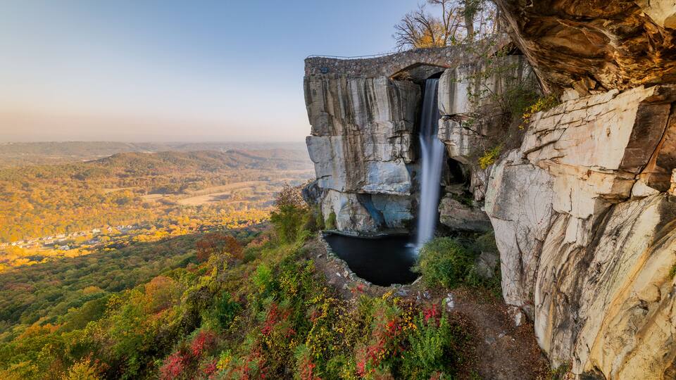 A waterfall cascading down a rocky cliff, surrounded by autumn-colored foliage and a valley below.