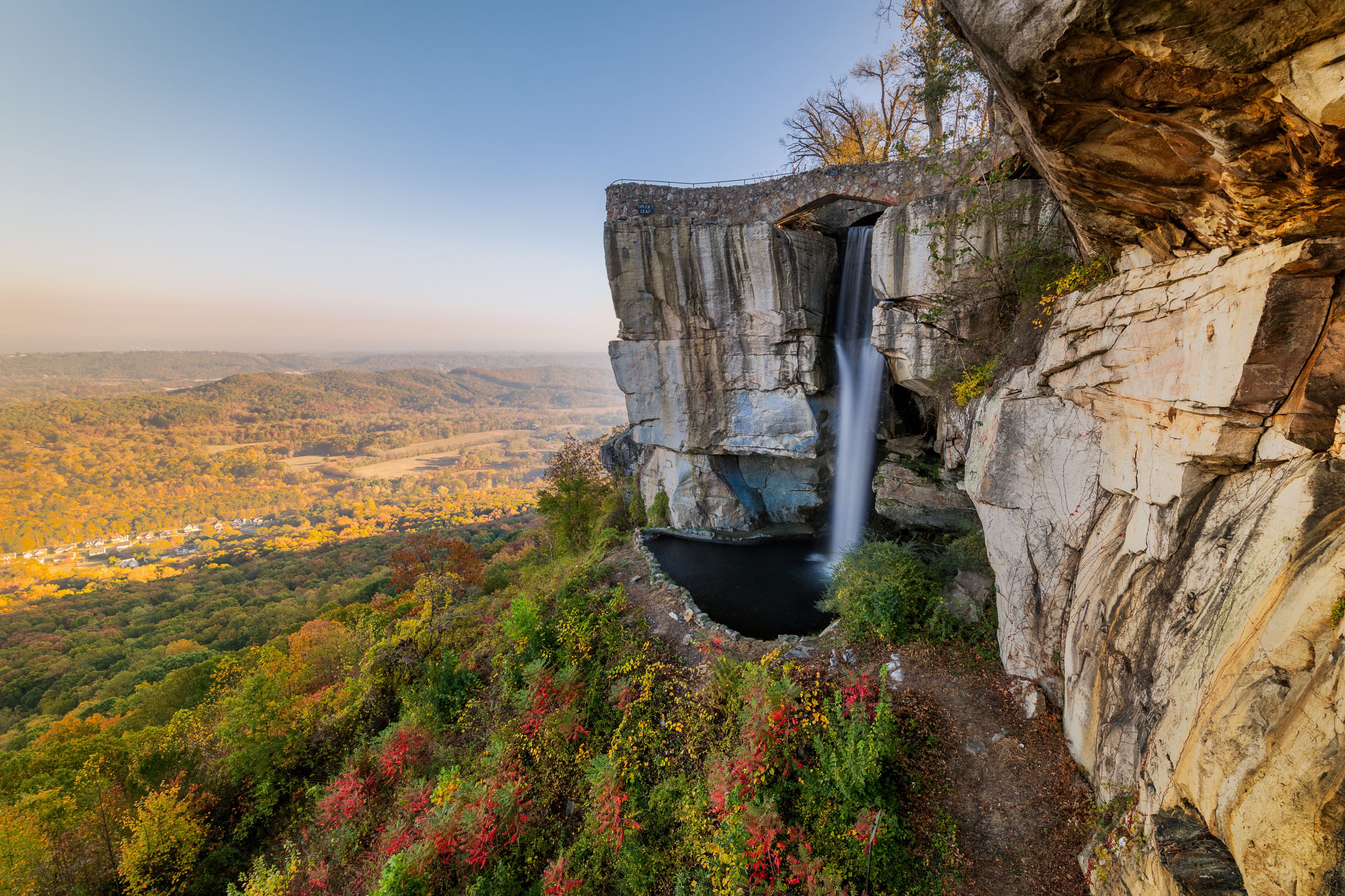 A waterfall cascading down a rocky cliff, surrounded by autumn-colored foliage and a valley below.