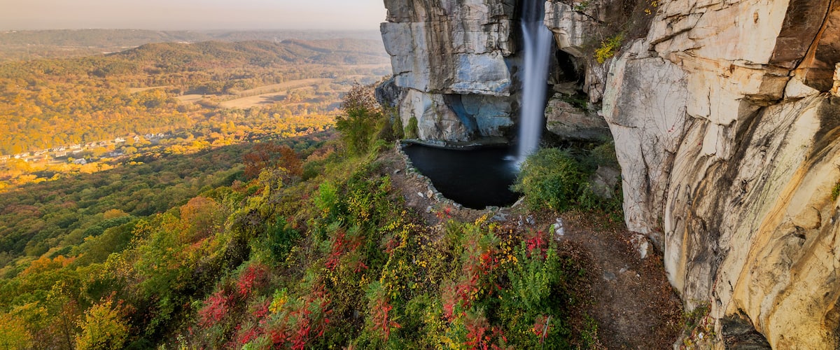 A waterfall cascading down a rocky cliff, surrounded by autumn-colored foliage and a valley below.