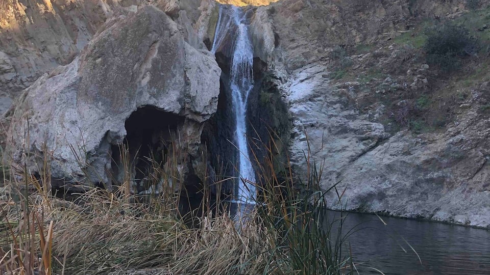 A waterfall cascading down a rocky cliff into a calm pool surrounded by dry grass and sparse vegetation.