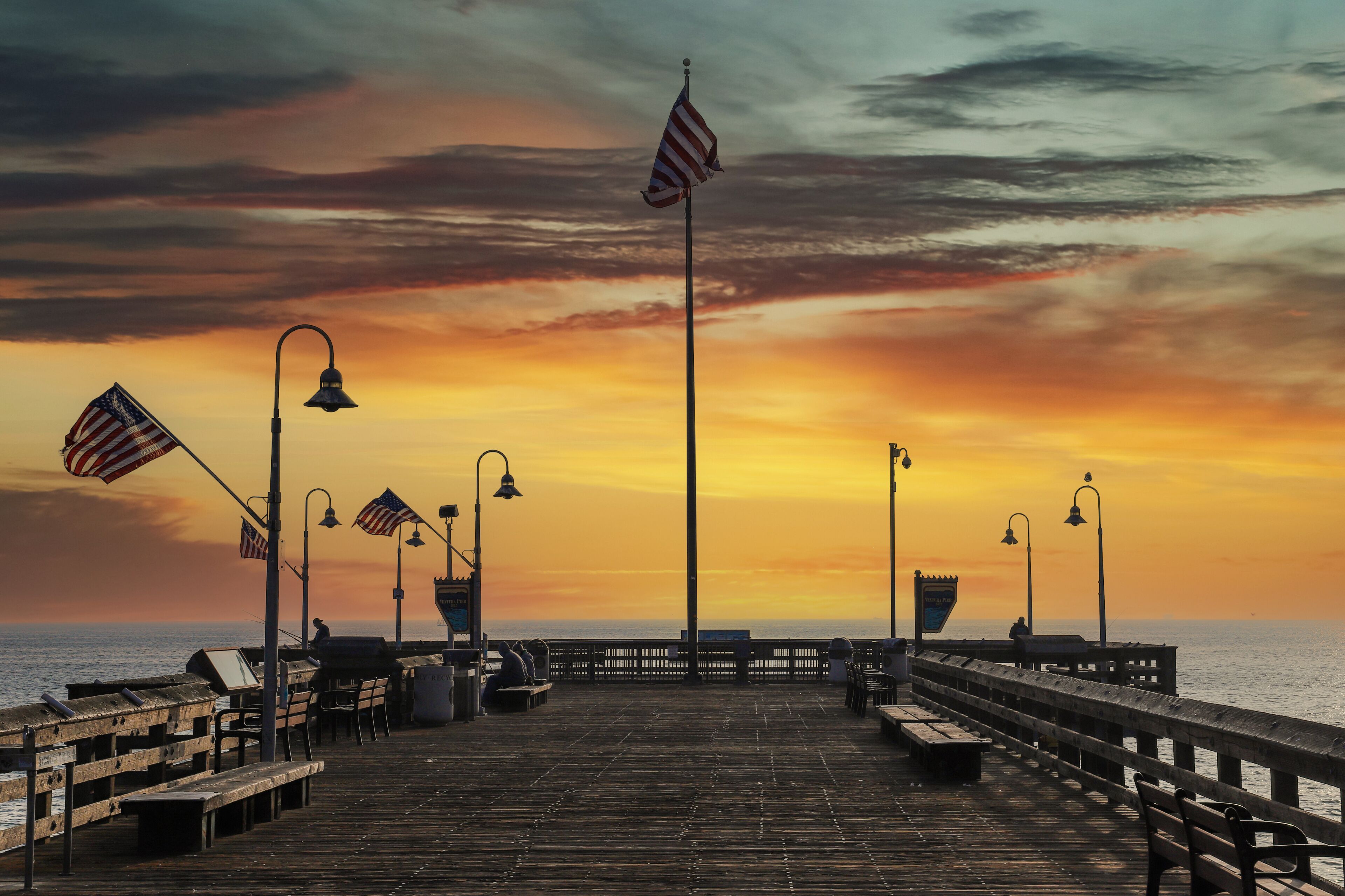 a shot of a long brown wooden pier with American flag flying on curved light posts with people fishing on the edge of the pier at sunset with powerful clouds at Ventura Pier in Ventura California USA