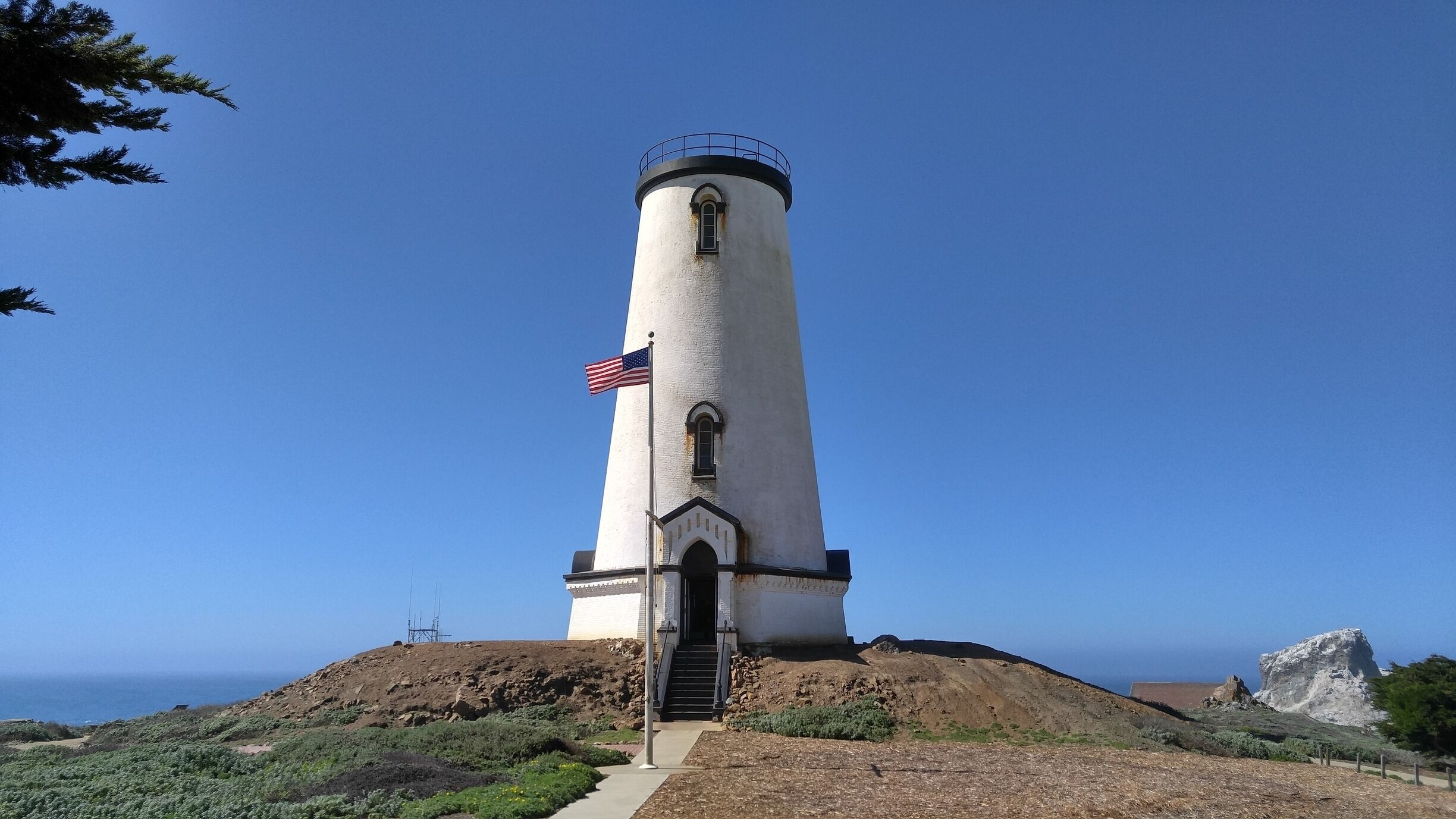 A tall white lighthouse with a flag on top, surrounded by hills and greenery.
